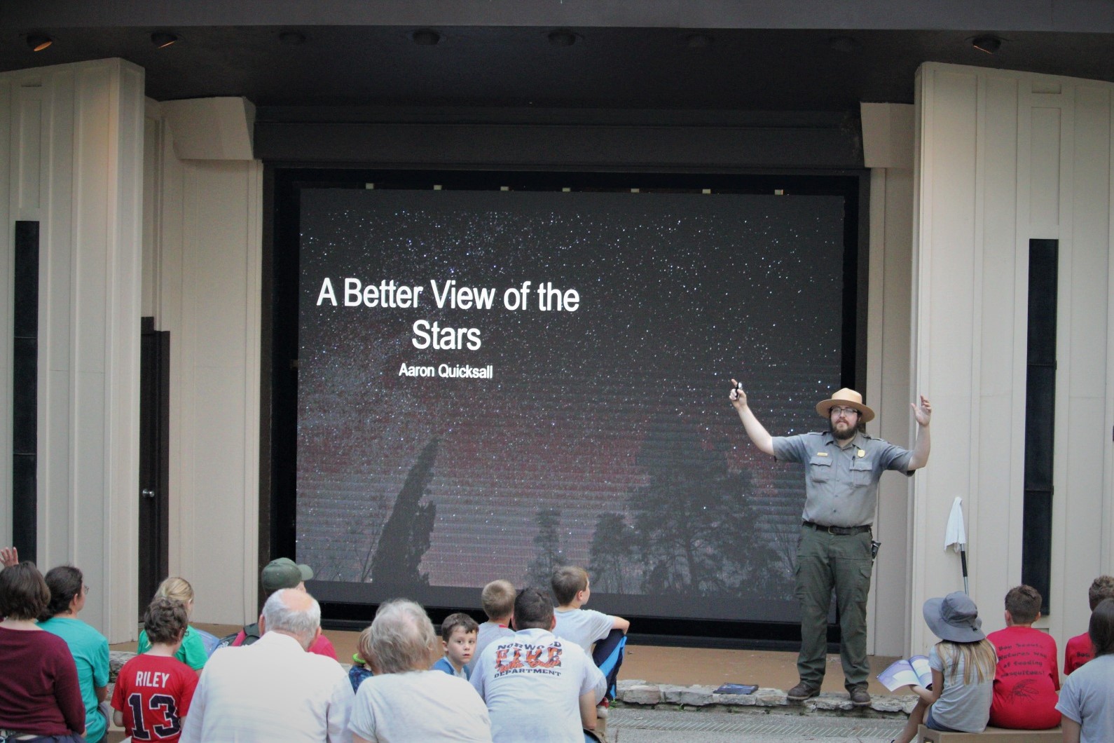 A person in a green and gray park service uniform stands in front of a crowd of seated people in an amphitheater.