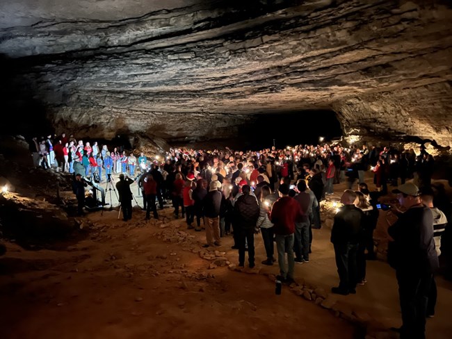 A large crowd of people hold candles and watch a choir singing inside a large and dark rocky room.