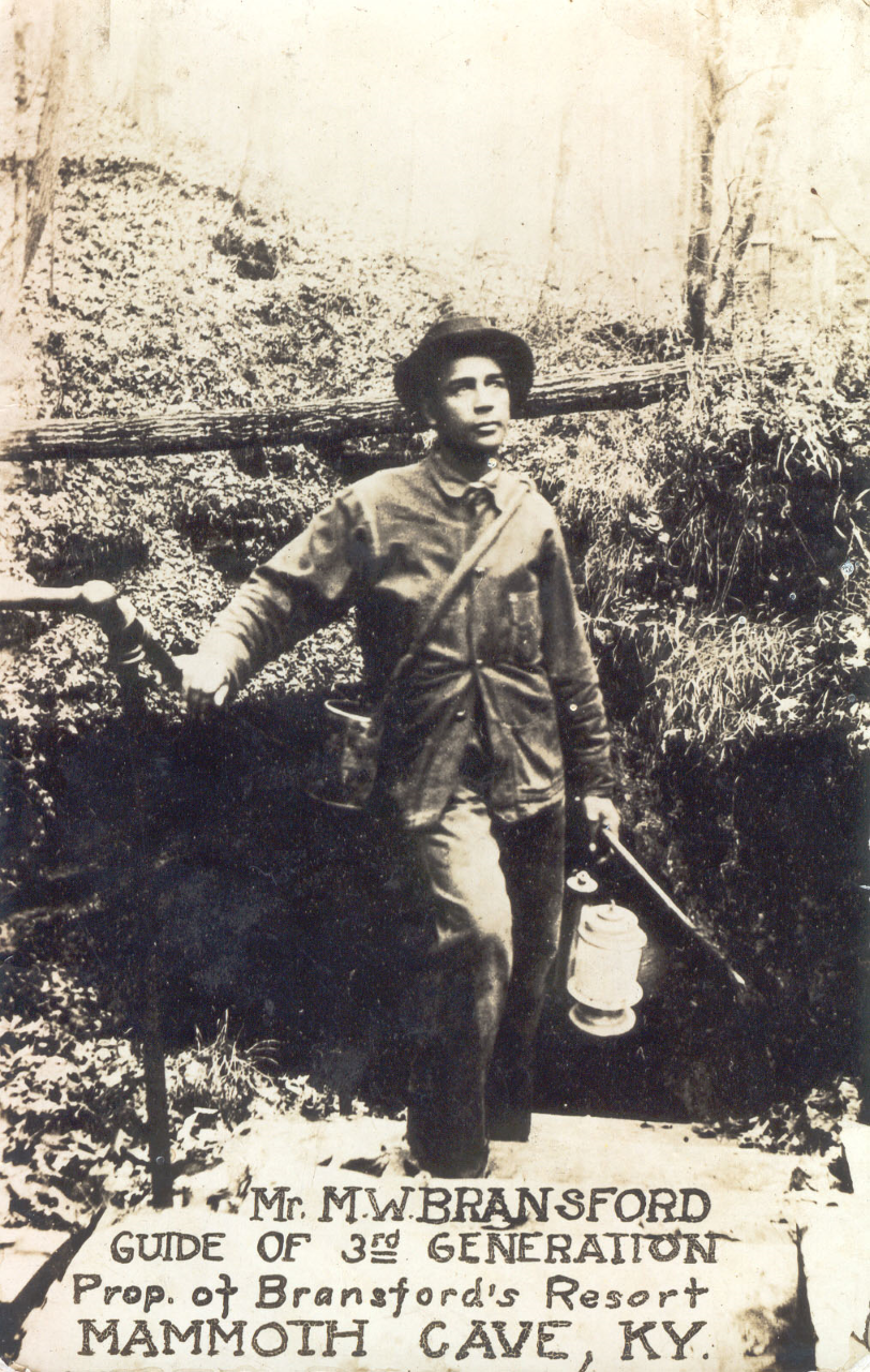 Historic black and white photograph of man in a coat carrying a lantern in front of a cave entrance. The words "Mr. M.W. Bransford Guide of 3rd Generation, prop. of Bransford's Resort, Mammoth Cave, KY" are written at the bottom.