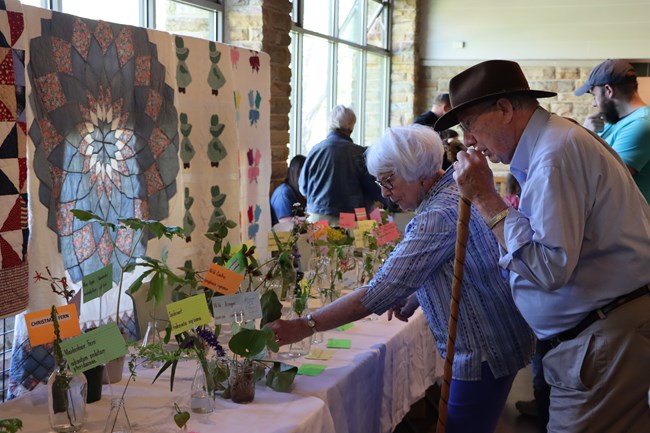People look at various flowers in glass containers on a table with colorful quilts displayed in the background.