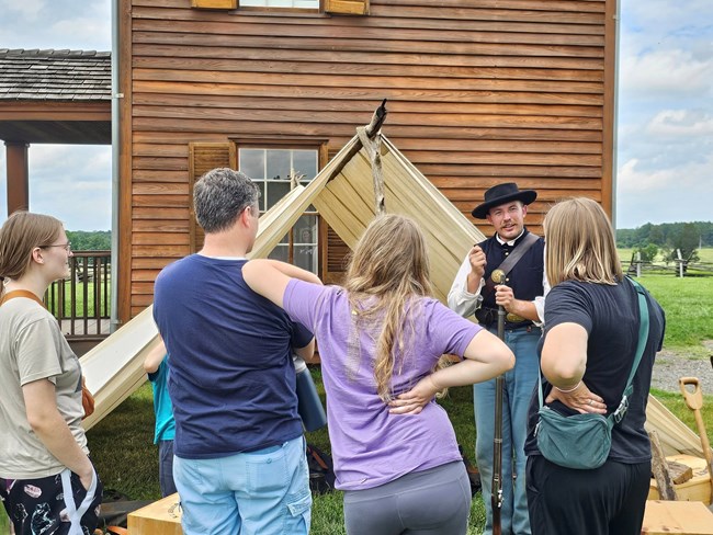 A ranger in Civil War Federal Uniform demonstrates soldier life to members of the community in front of a tent.
