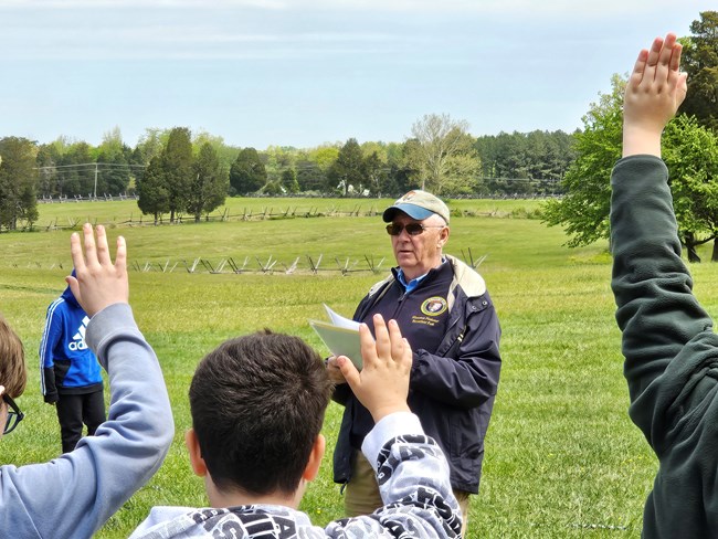 Students raising their hands on Henry Hill during an education program given by a volunteer wearing his uniform, navy blue jacket and tan hat, and holding papers.