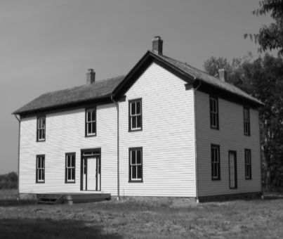 Black and white photo of a white wood house with ten windows, two doors, and three chimneys shown. The yard is grass with trees in the background.