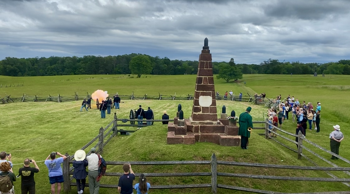 A crowd of visitors gather around a cannon firing demonstration and monument.