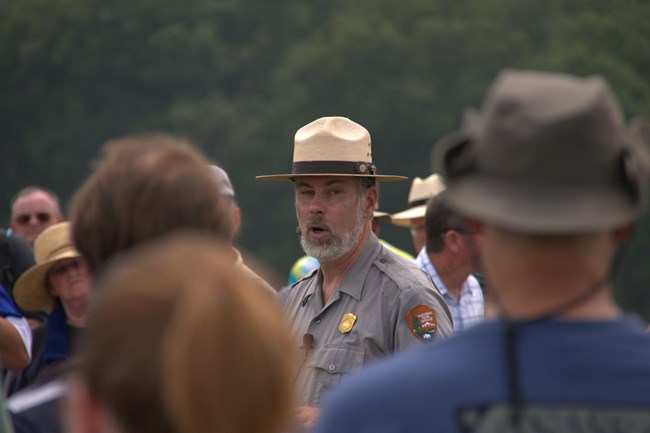 A park ranger leads a tour with a group of people around him, from the shoulders up. The landscape is green behind them.