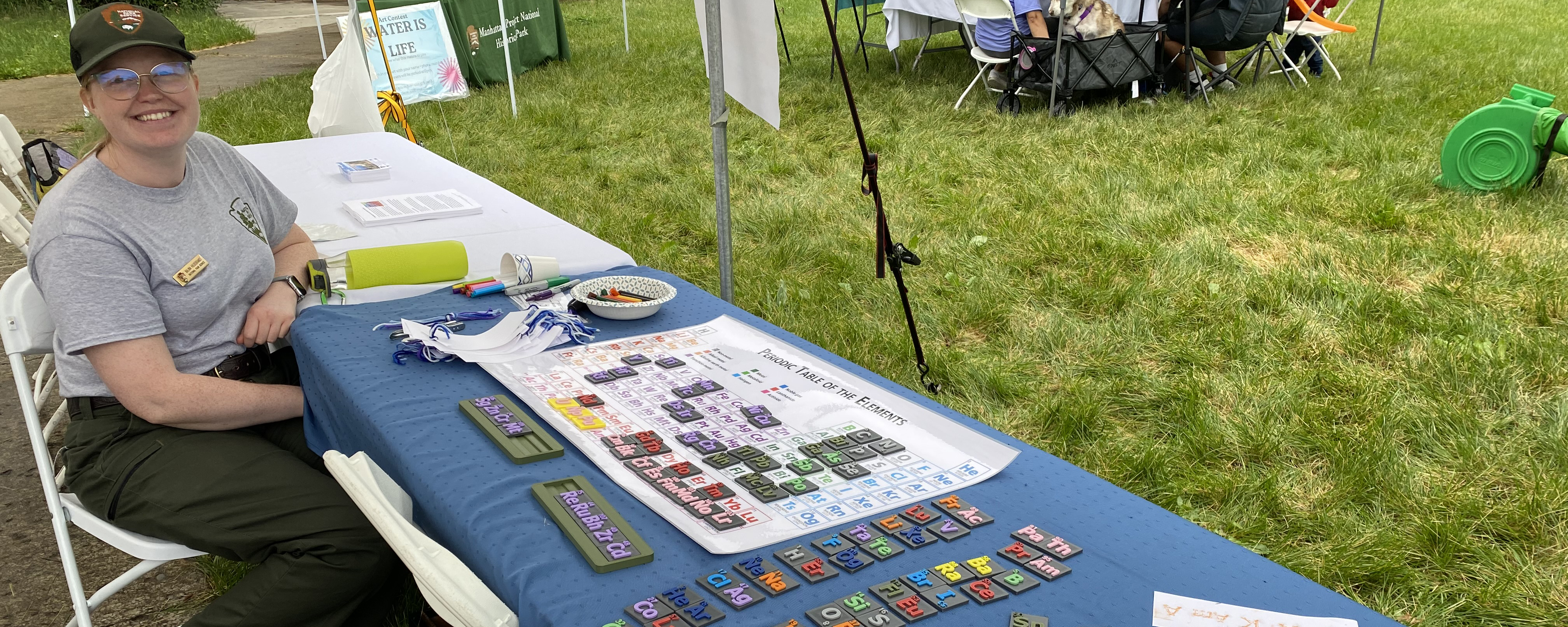 A woman sits at a table with many small plastic squares and a periodic table of elements poster.