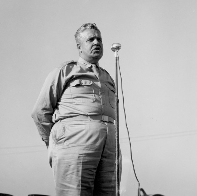 General Leslie Groves, chief administrator of the Manhattan Project, stands before a stand mic giving a speech.