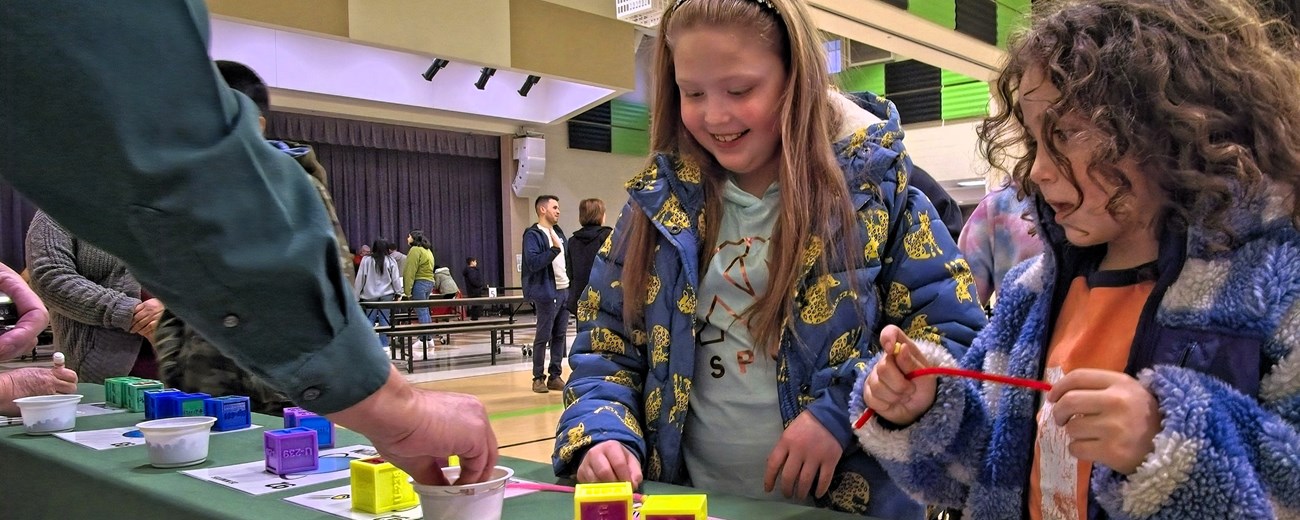 Two girls stand facing a table playing a game while a man behind the table explains the game.