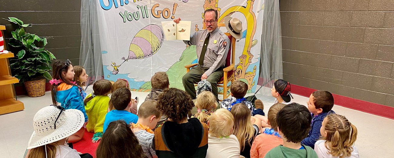 A seated uniformed man holds high an open book, reading to 20 kids seated on the floor in front.