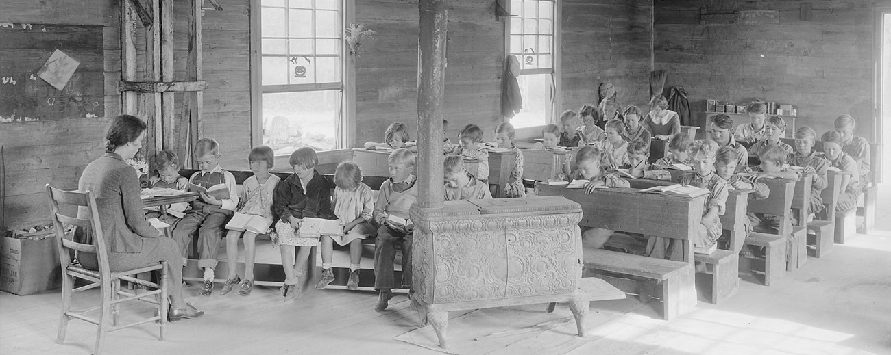 A small primitive classroom, with children seated at pews and a female teacher seated at the front of the class. A large wood burning stove separates the students from the teacher.
