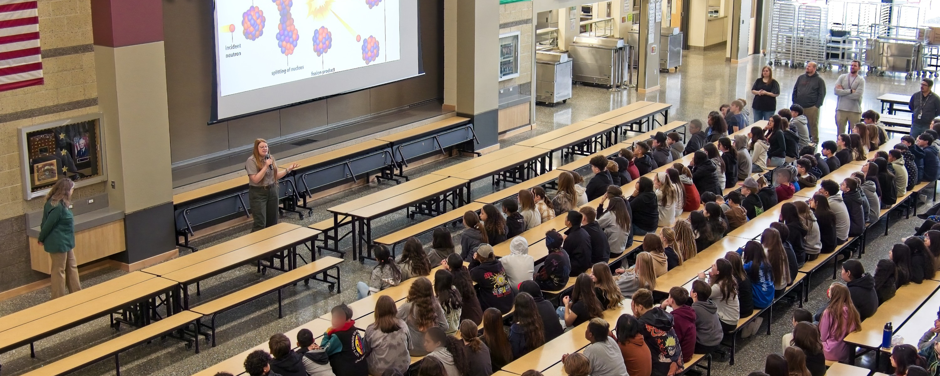 200 students sit at long tables looking at a woman wearing green pants and grey shirt talking in front of a large screen.