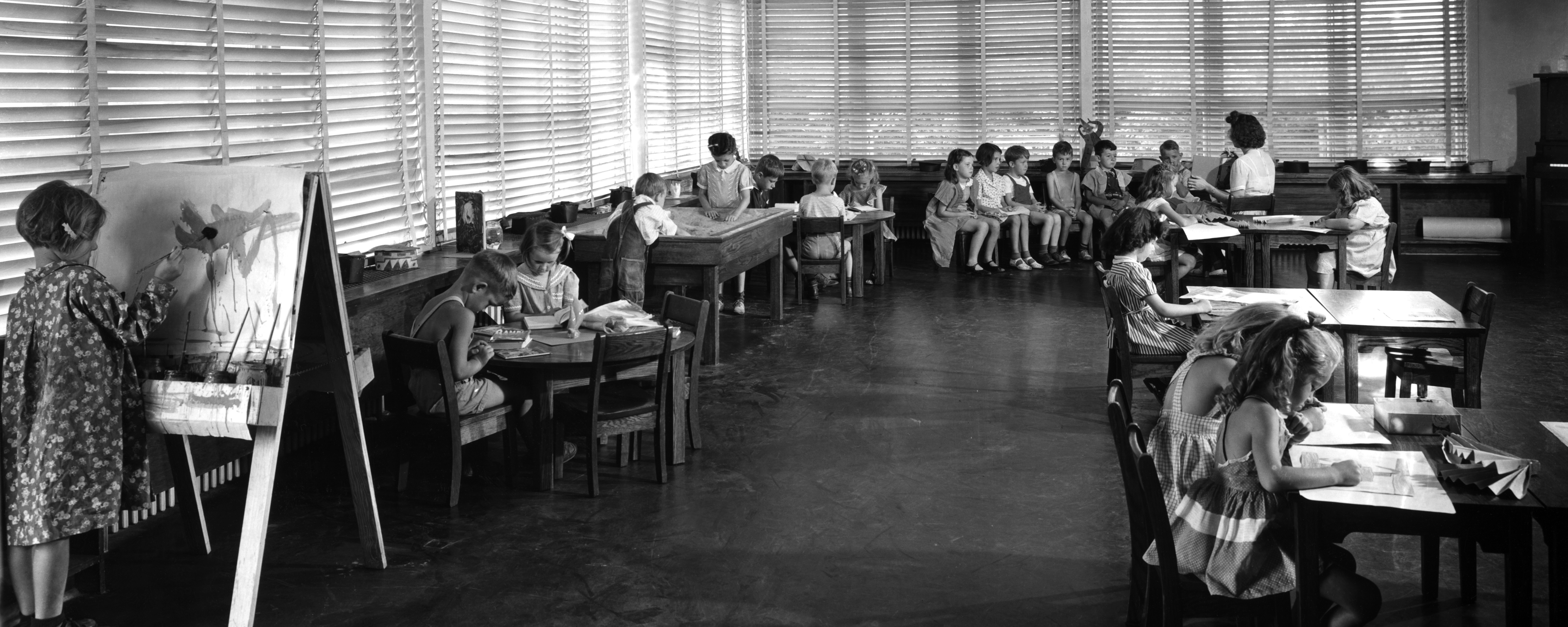 A classroom scene from 1944. 1 child stands at easel painting; groups of children sit at four tables; a teacher reads to a group of six in the back of the room.