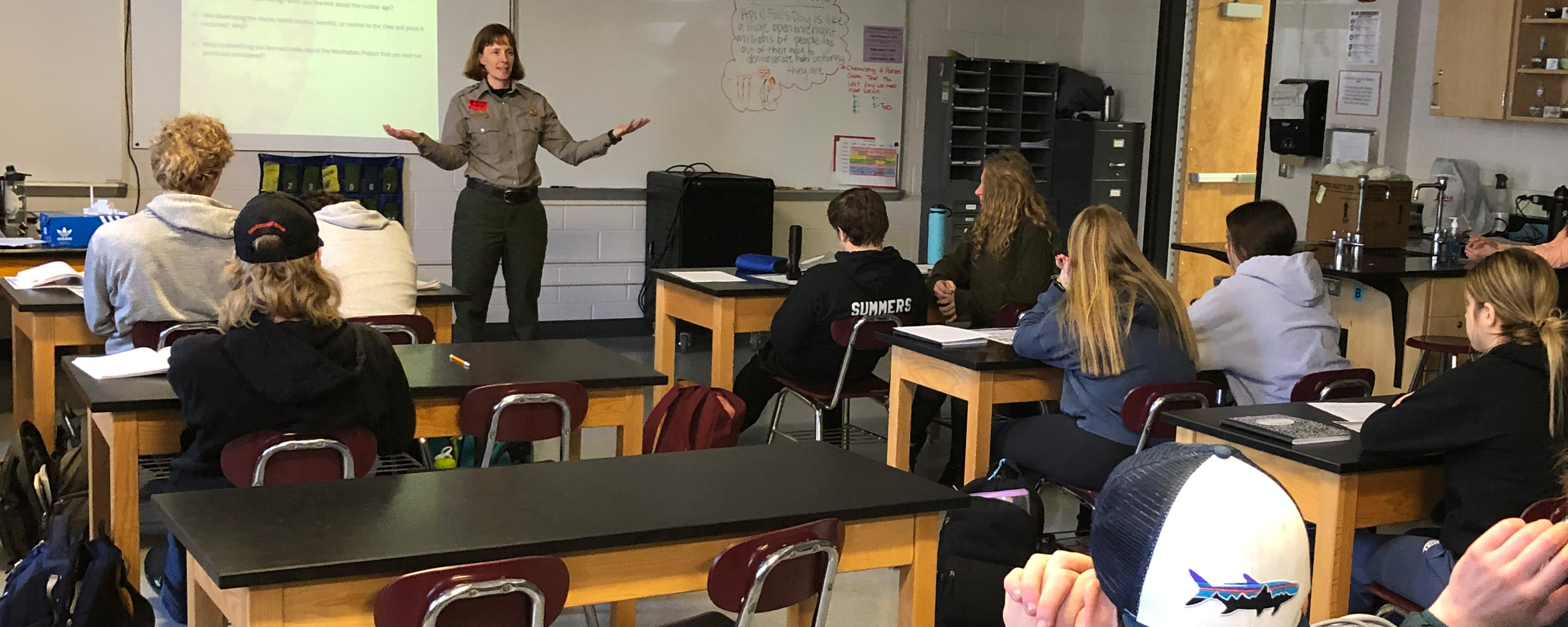 A women in a gray shirt and green pants ranger talks in front of 9 students sitting at desks.