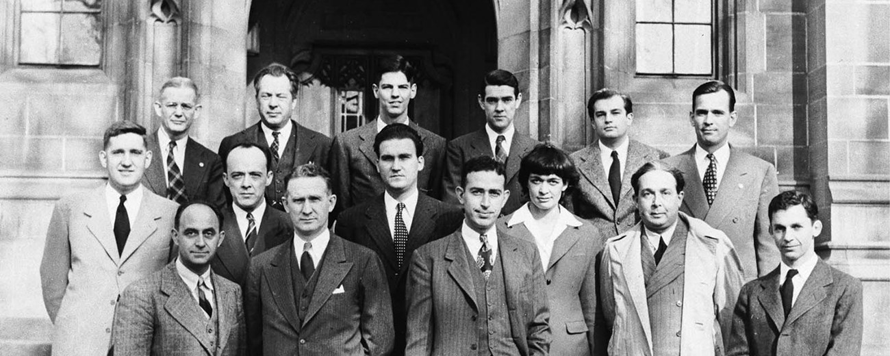 Fourteen men and one woman stand at the entrance of a large university building.
