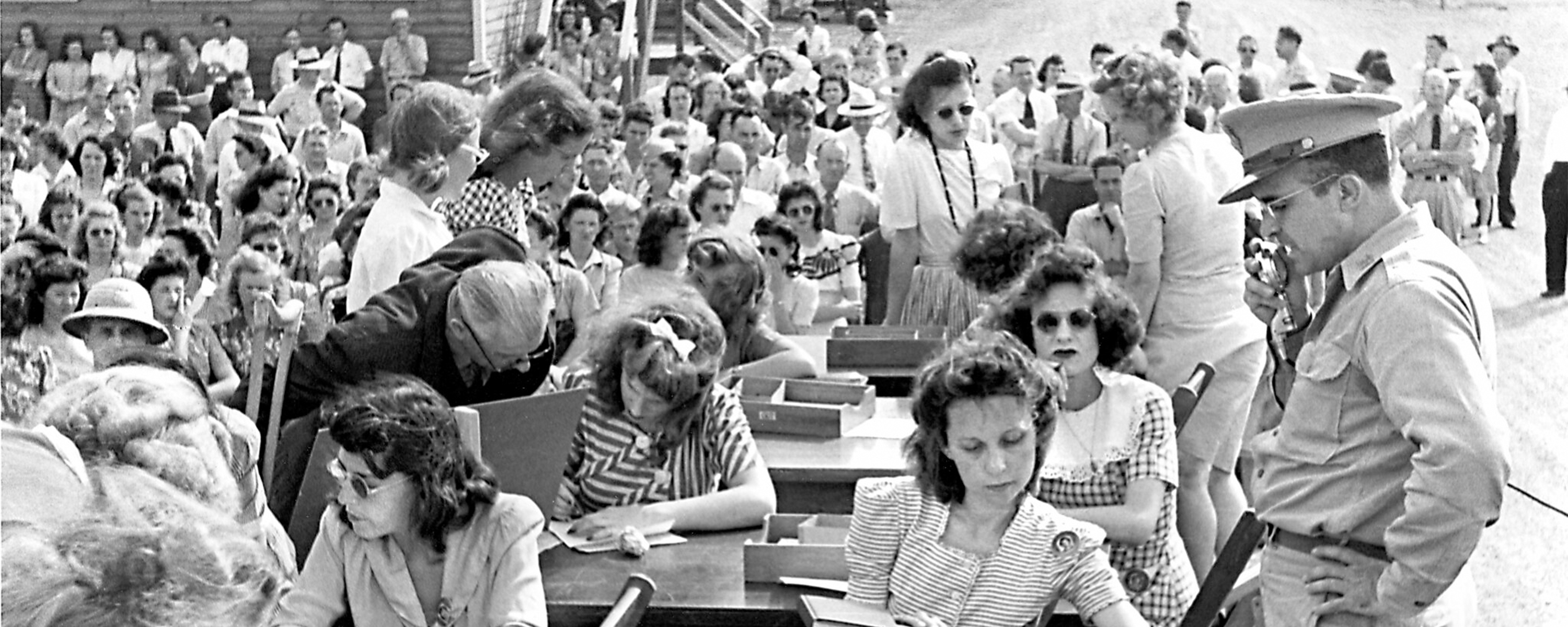Several men and women seated at outdoor desks with military personnel looking on.