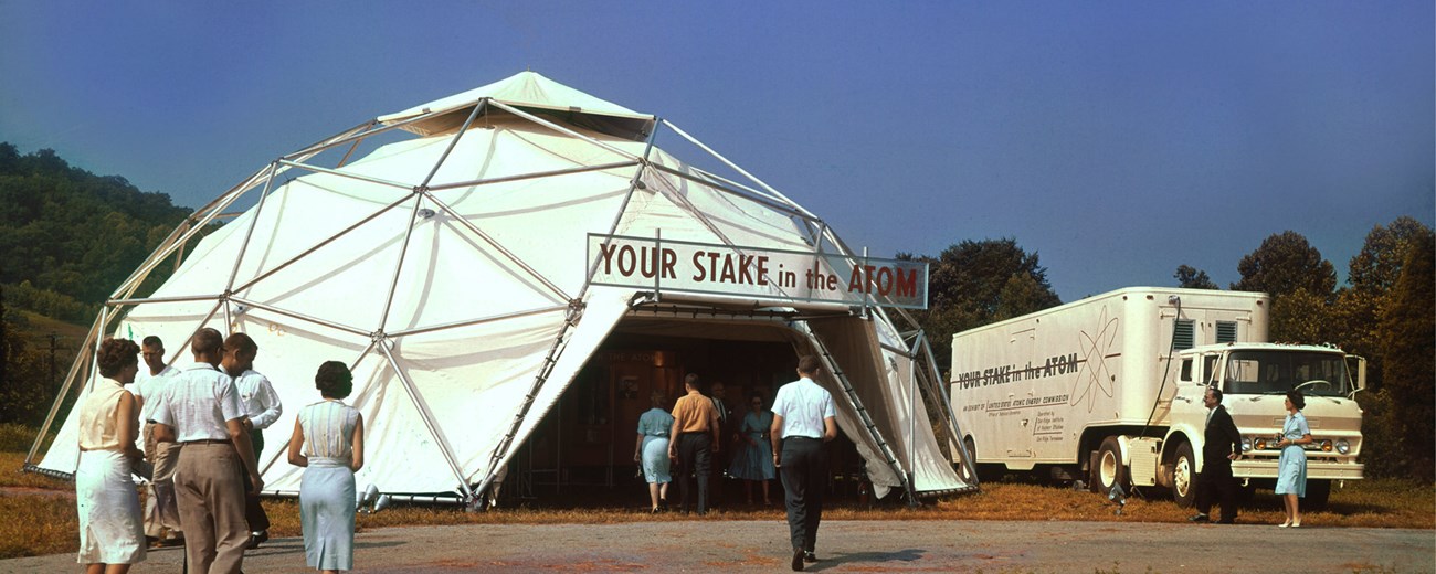 Several people walk toward a geodesic dome with a sign that reads: “Your Stake in the Atom.”