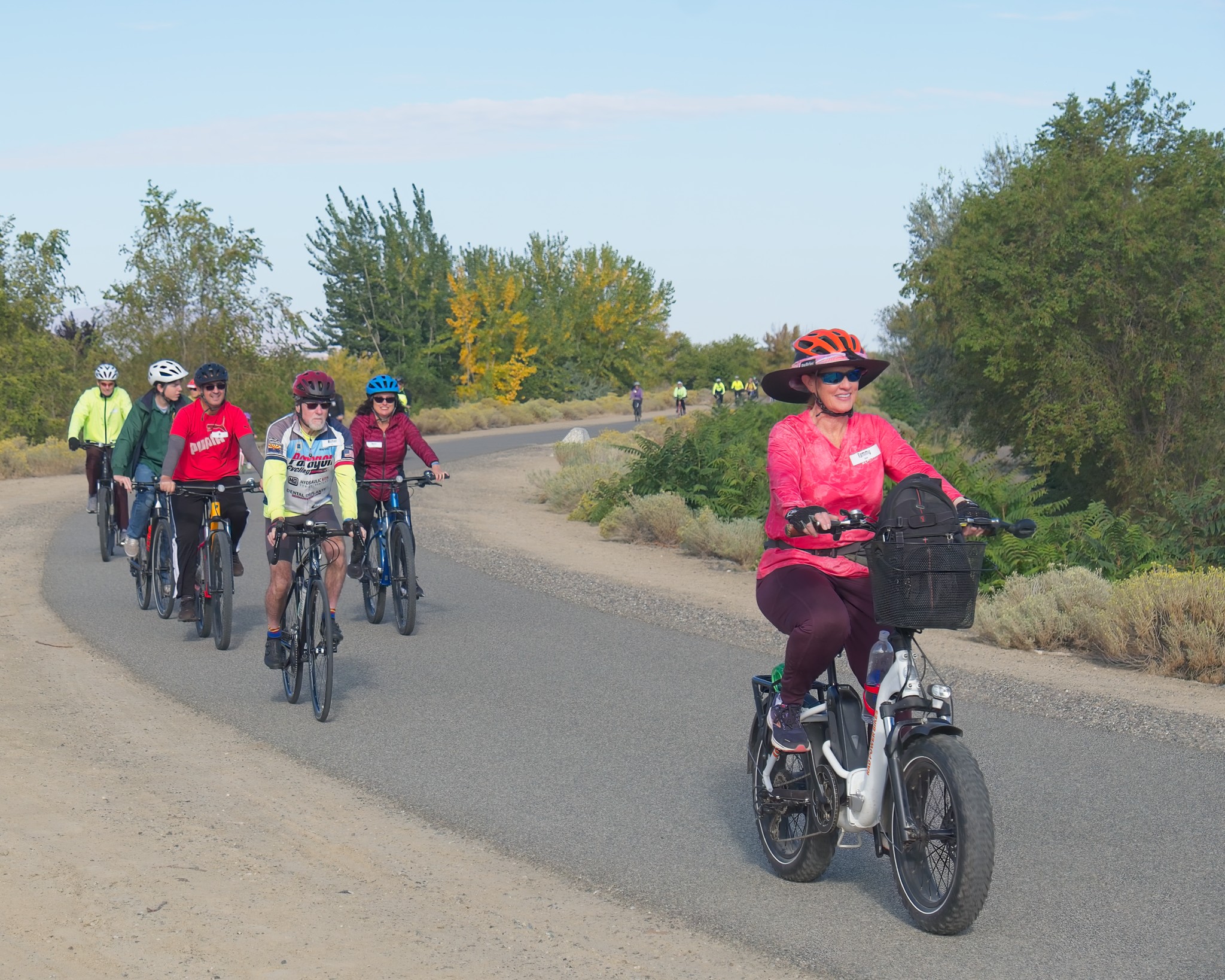 5 cyclists on a pave path ride towards the camera on a bright sunny day. Trees with green and yellow leaves line the path and with a group of 10 riders are seen in the background.