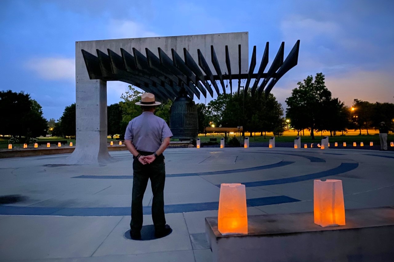 A man stands with arms behind his back facing a large bell hung from a sturdy concrete structure. Lunch bags illuminated with candles line the walkway.