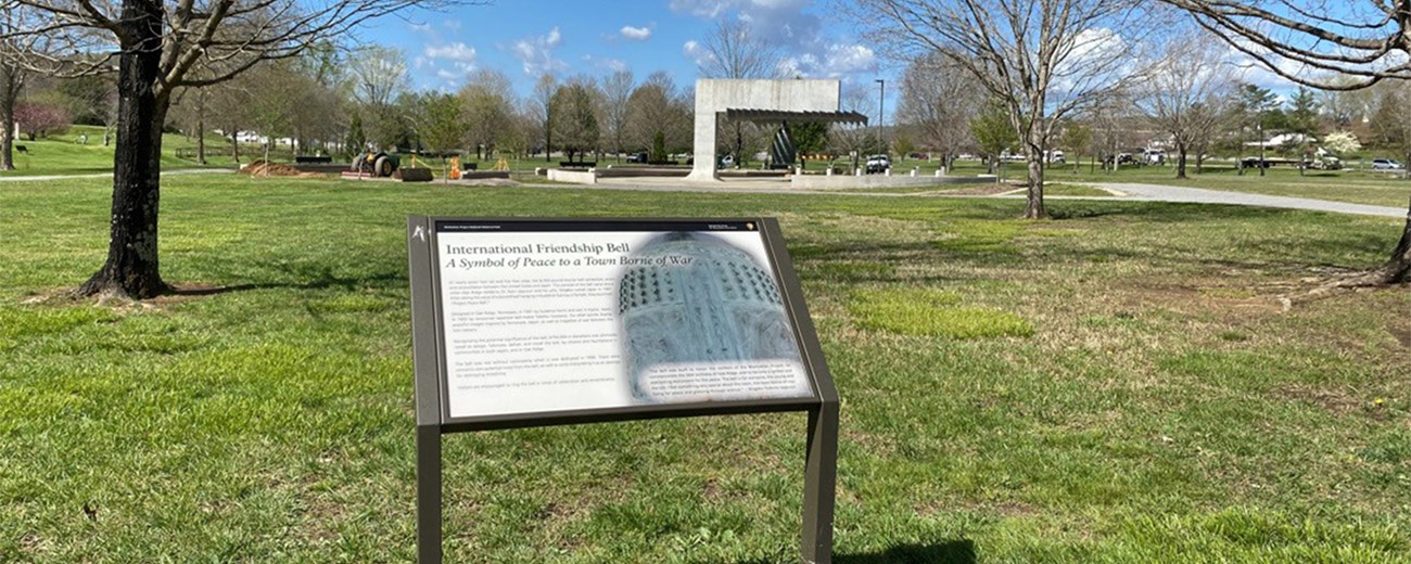 A wayside exhibit in a city park with a large bronze bell in the background.
