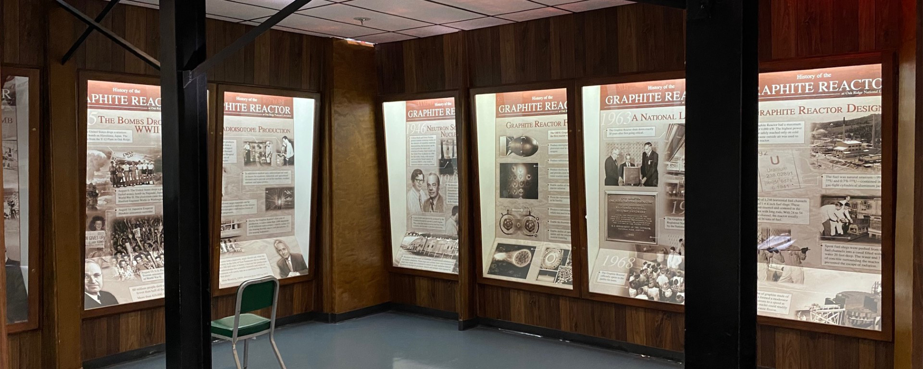 A small wood-paneled exhibit room with illuminated interpretive panels along the walls. A small green chair sits next to a support pillar in the center of the room.