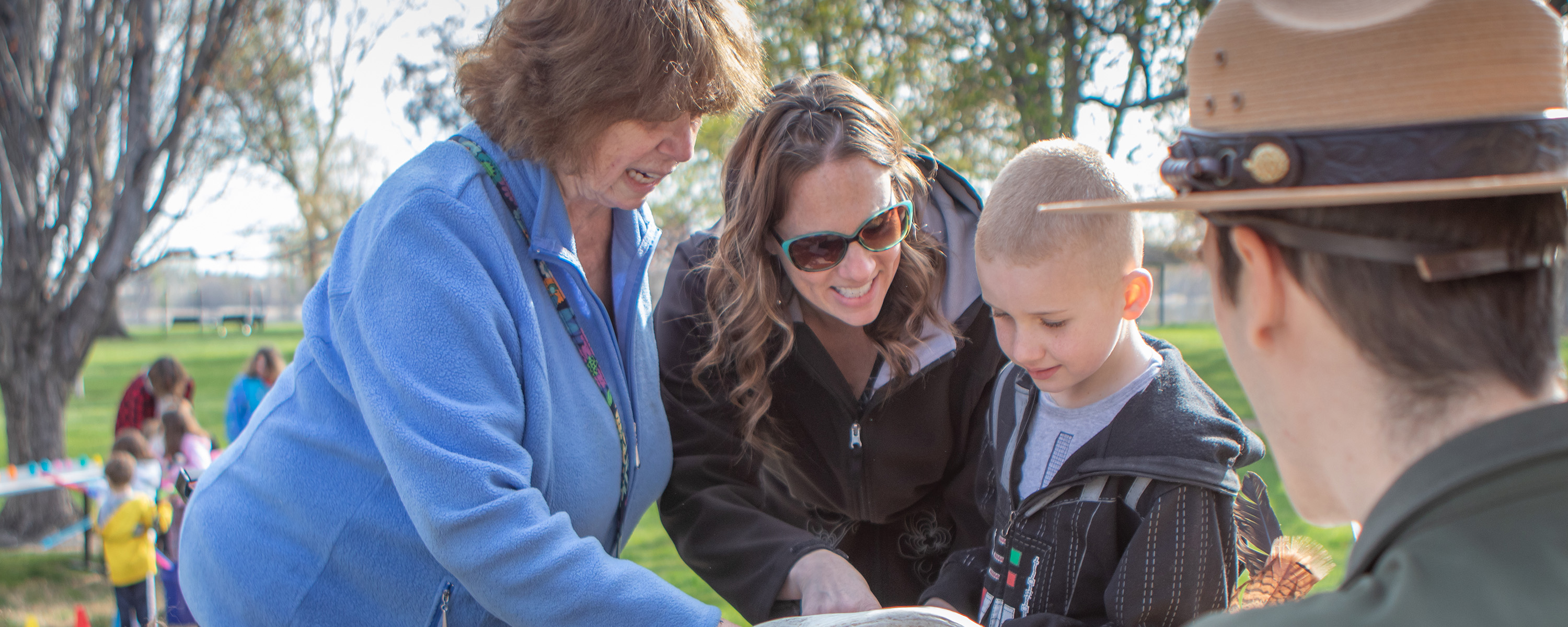 A young child looks at a display table hosted by a uniformed park ranger while two older women look down and point out something to the child.
