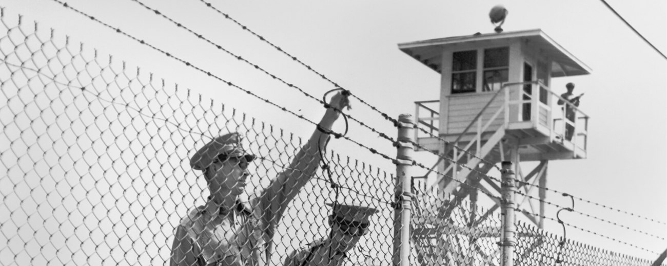 Two men in police uniforms inspect a barbed wire fence with a guard tower behind them.