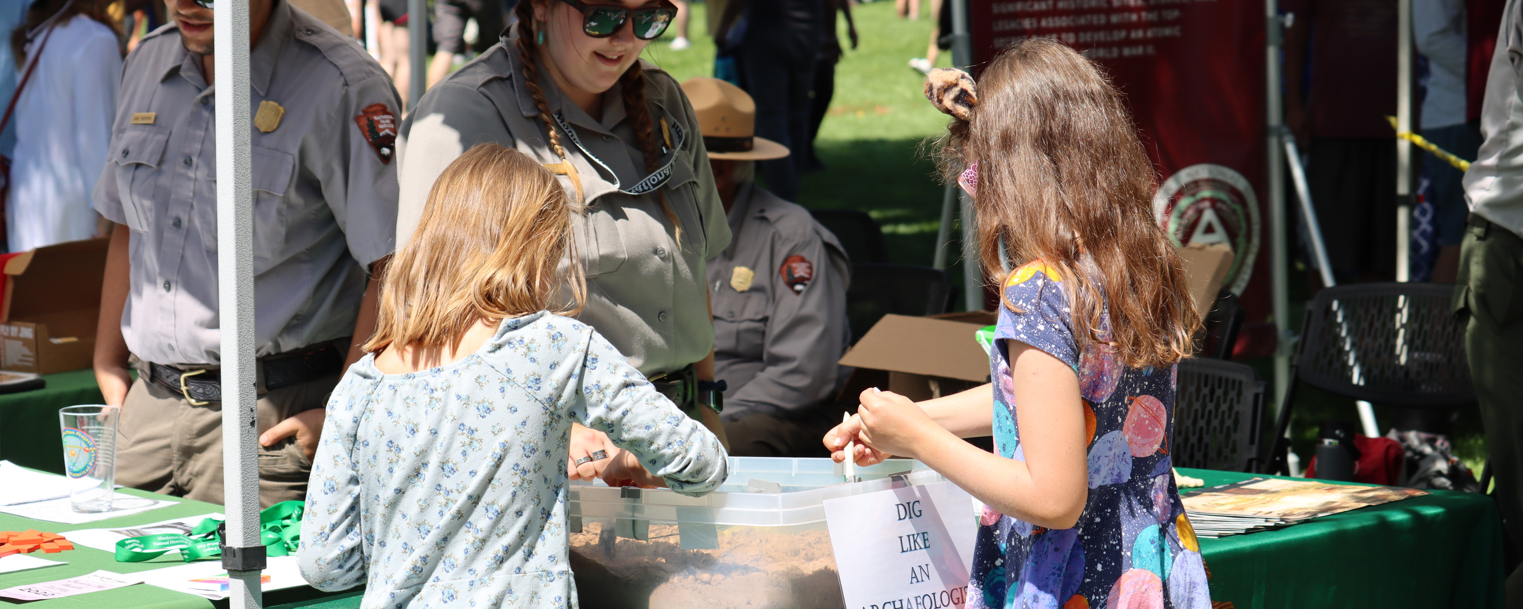 A uniformed ranger watches and smiles as two young girls play with items on a table.