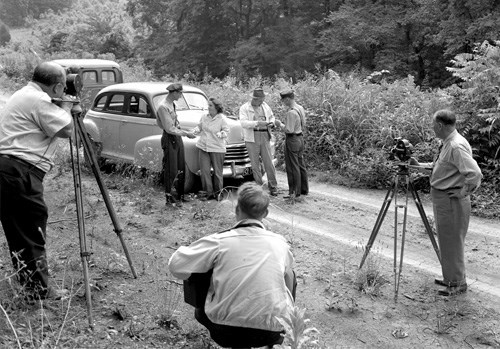 A black and white photo of a group of people with tripods set up and posing for photos in front of a car.
