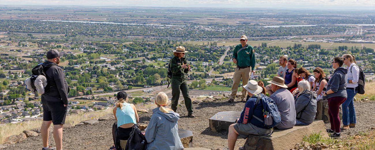 A group of people sit and stand around two people in uniforms talking at the top of a hill with a view of the town below in the background.