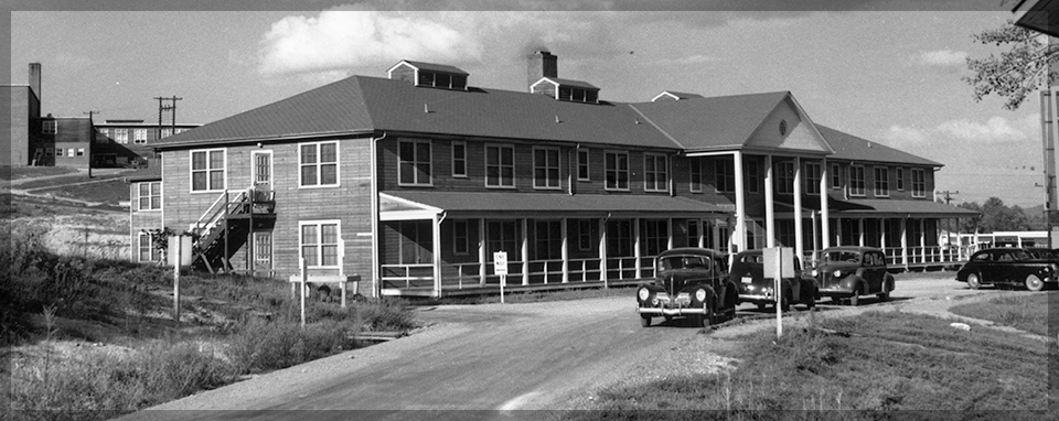 Black and white photo of a building with many windows and at least two stories. There are cars parked on a road in front of it.