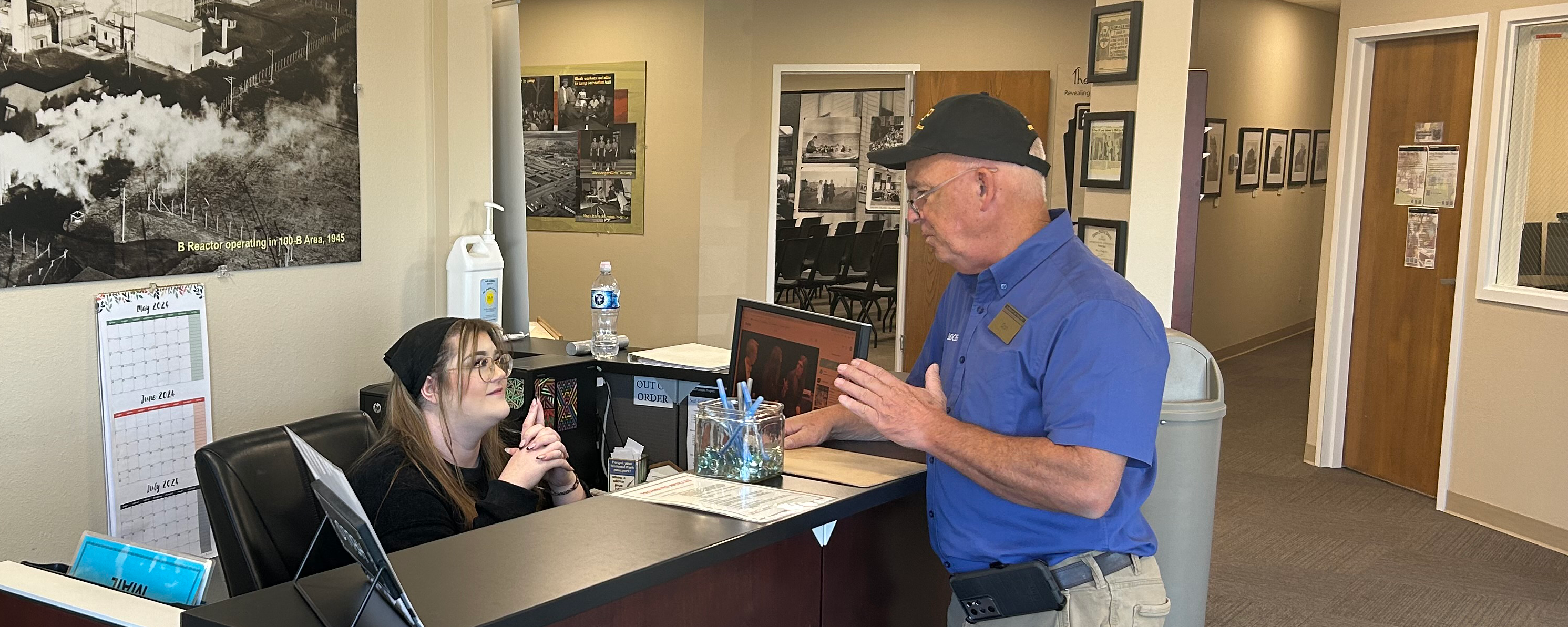 Man in blue shirt stands at visitor center desk, staff sits attentively behind desk. Doors and black and white photos line walls in the background.