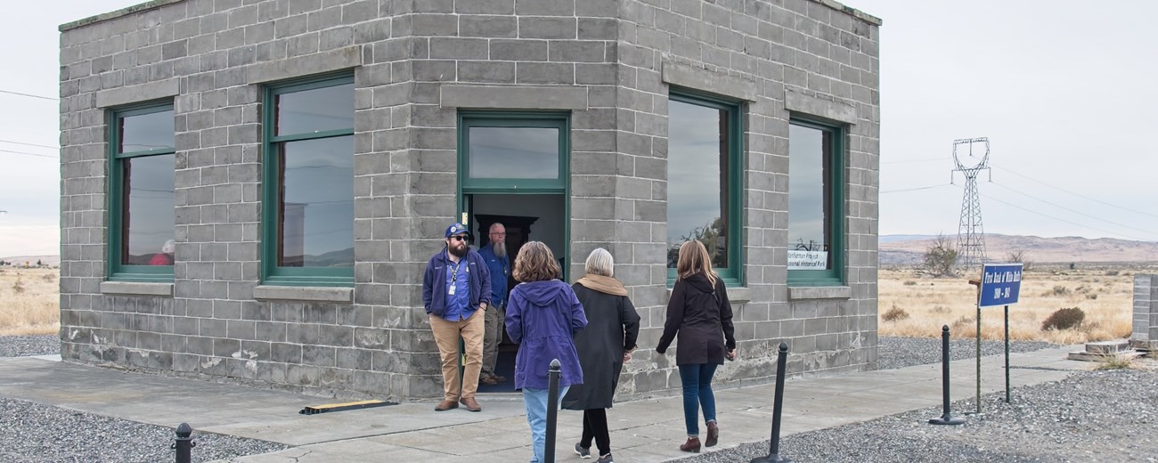 A walking tour heads back towards a gray stone block building with four large windows and a single glass door under cloudy skies.