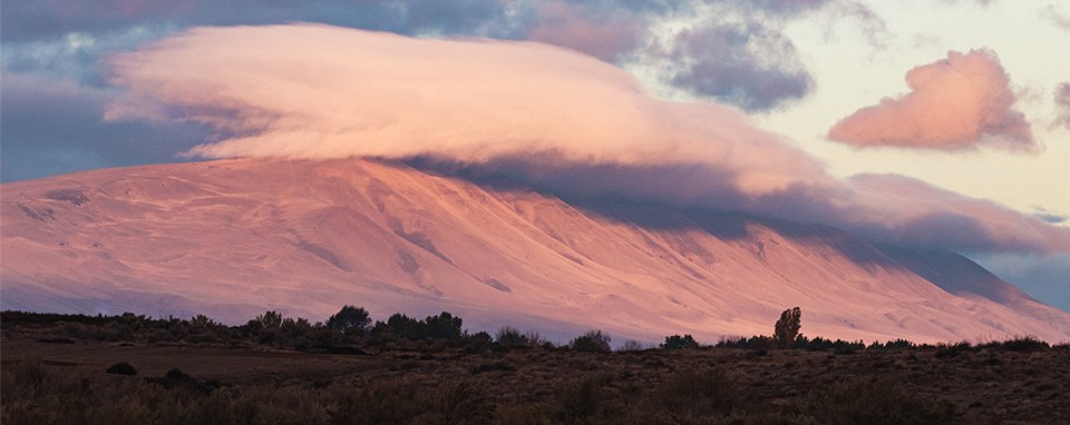 A color photo of a mountain bathed in pink light with a pink cloud above it. The foreground is treeless but dark.