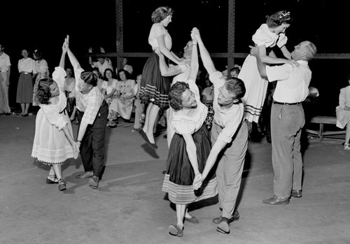 A black and white photo of 3 couples dancing with onlookers in the background.