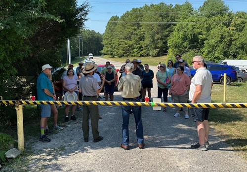 A group of people stand behind a yellow and black striped gate watching a uniformed ranger speak.