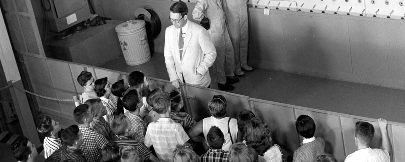 A man in a light colored suit stands in front of a reactor face while several children look at him from the floor below.