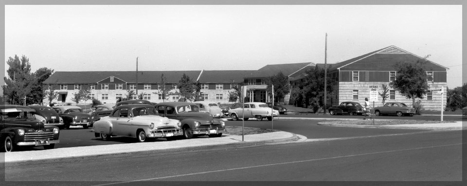 A black and white photo of large building with many windows. There is a parking lot in front full of cars.