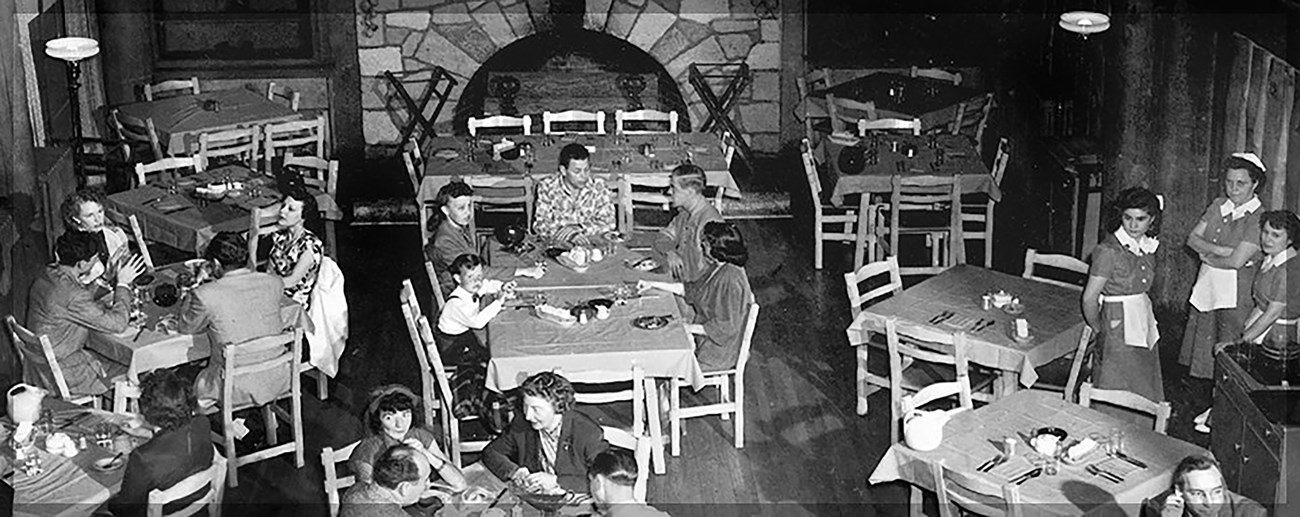 From above, a black and white photo of people eating at several square tables in a dining hall with a stone fireplace in the background. Three people in aprons are standing to one side.