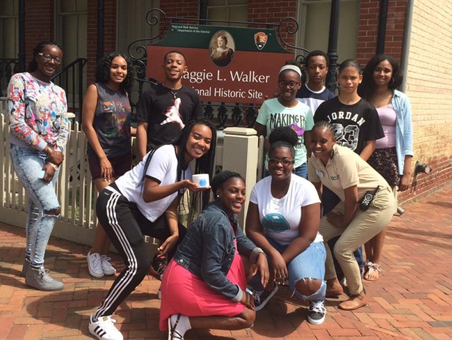 Student participants in the Summer Youth Leadership Institute pose with an intern in front of the Maggie L. Walker National Historic Site sign