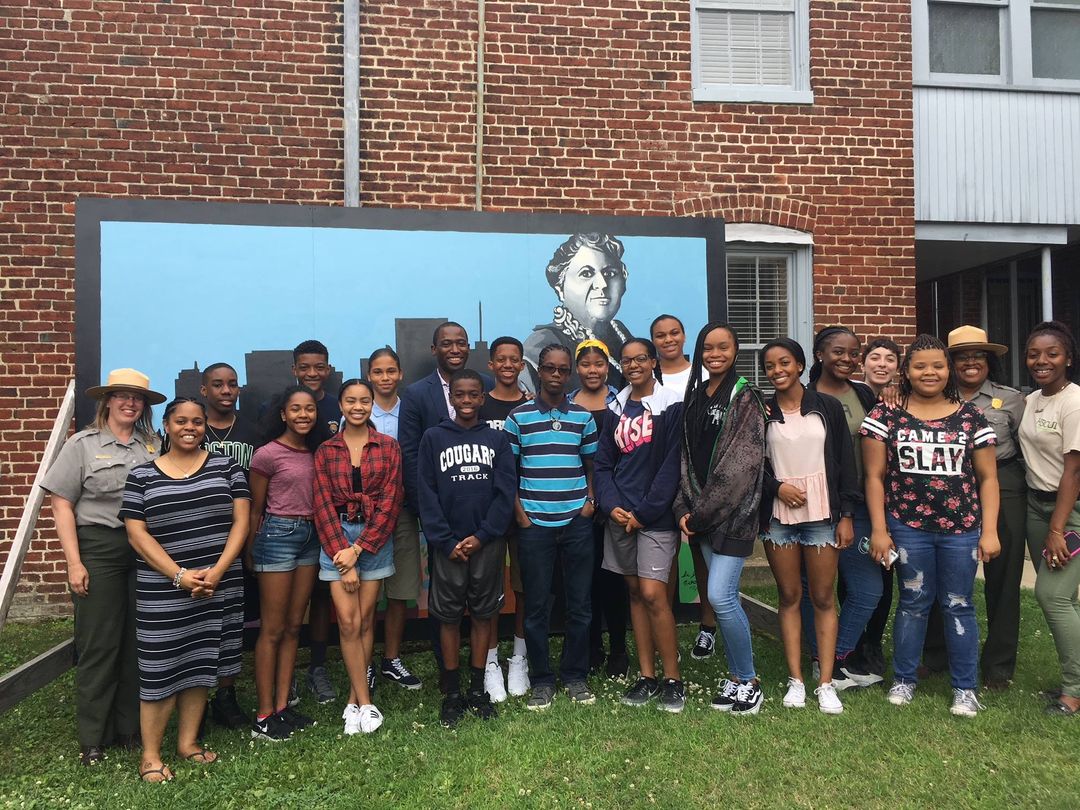 Students and Rangers pose in front of a mural of Maggie L. Walker in our courtyard