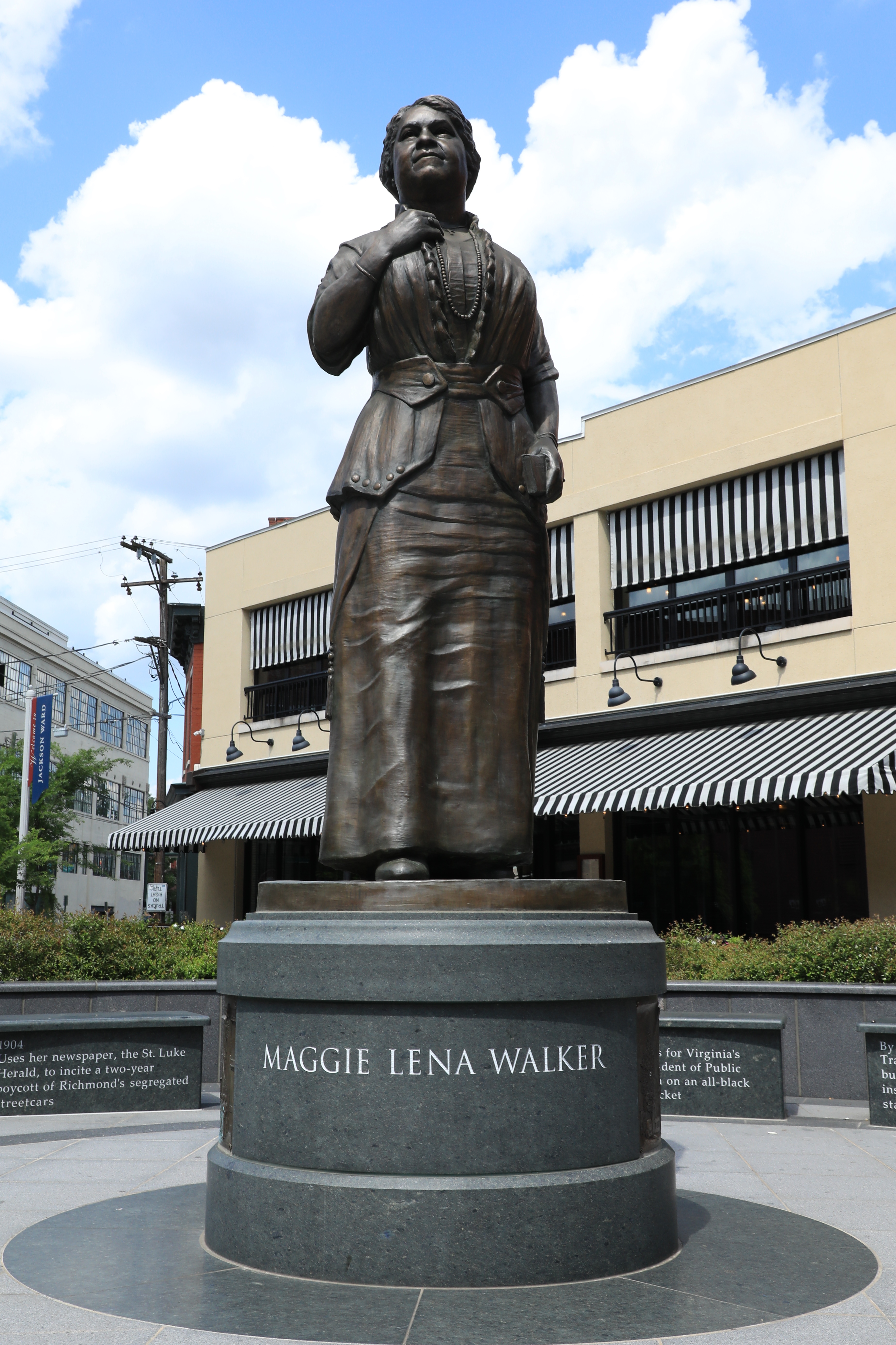 Frontal view of a bronze statue on a pedestal with buildings, blue sky, and clouds in the background
