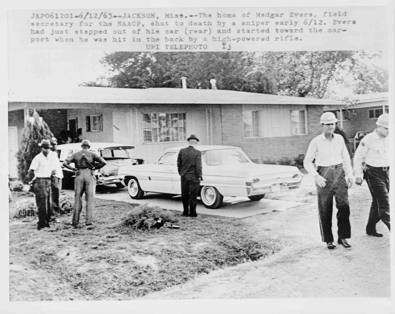 Historic photo of law enforcement officers and neighbors in Evers yard investigating the crime scene the day after the shooting. Typed label at top.