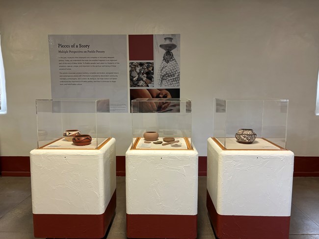 Three square display cases, dark red bottom, white middle third and  tall glass on top. These contain ancient and modern pottery. On the wall hangs a large exhibit about pottery