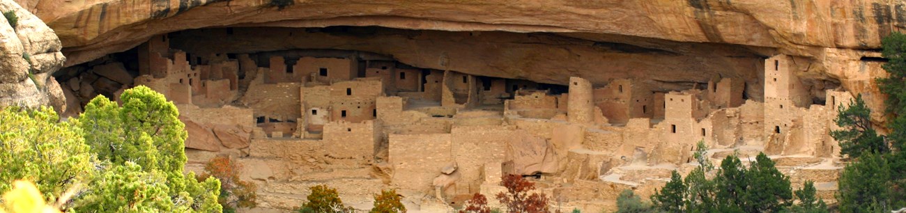 A panoramic view of a long Ancestral Pueblo cliff dwelling in setting sun light. It includes towers, kivas, dwellings, and storage areas.