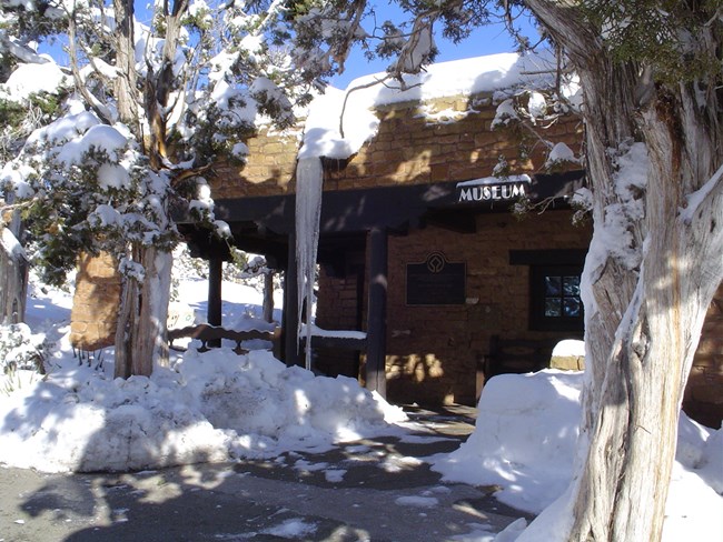 A brown museum sign hangs from a native sandstone building above a shoveled walk with snow covered trees