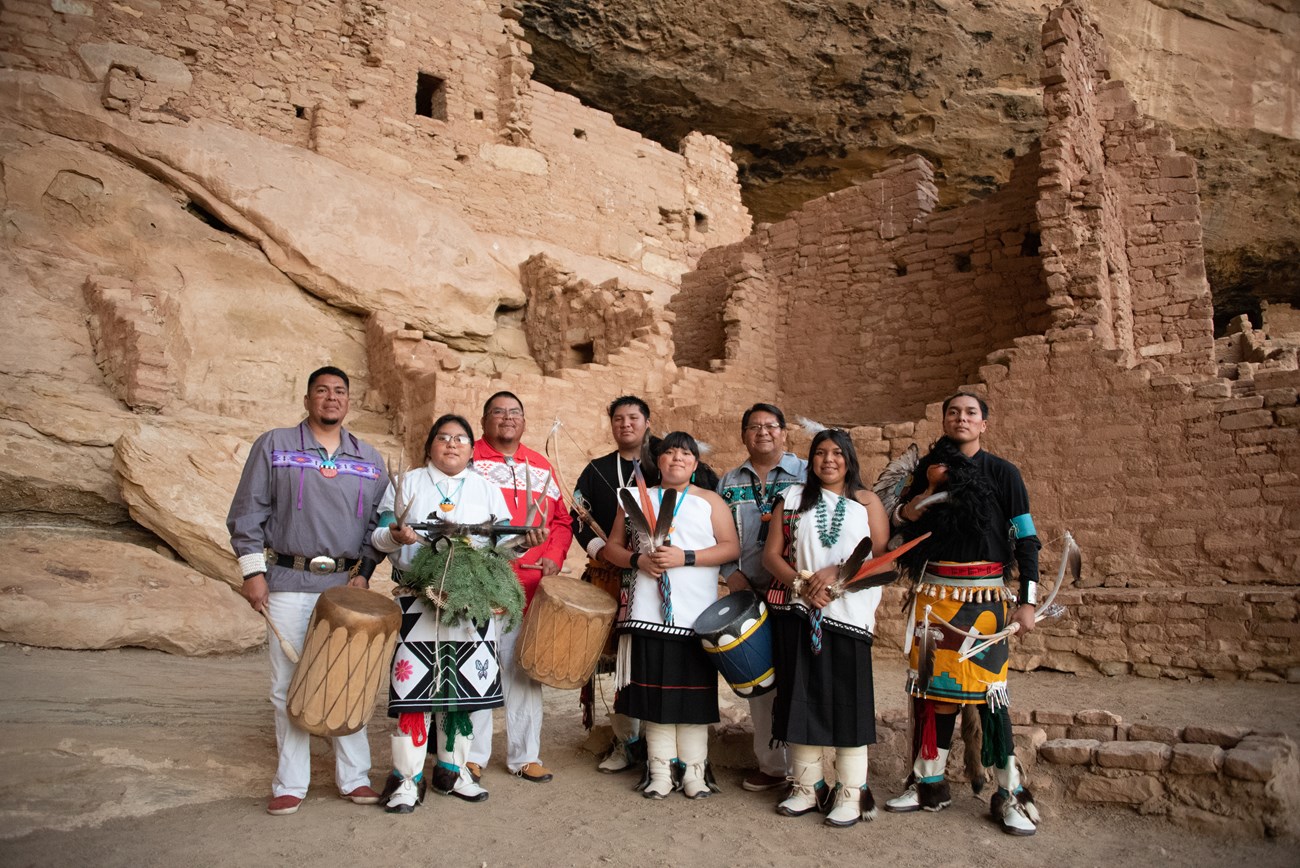 Eight Pueblo dances dressed in regalia stand in the plaza in front of an ancestral site