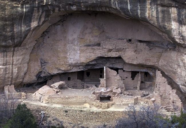 A two story ancestral dwelling built with a sandstone alcove. It includes four round kiva, broken stone walls with doors and windows. The surrounding sandstone is heavily striped with natural black varnish.