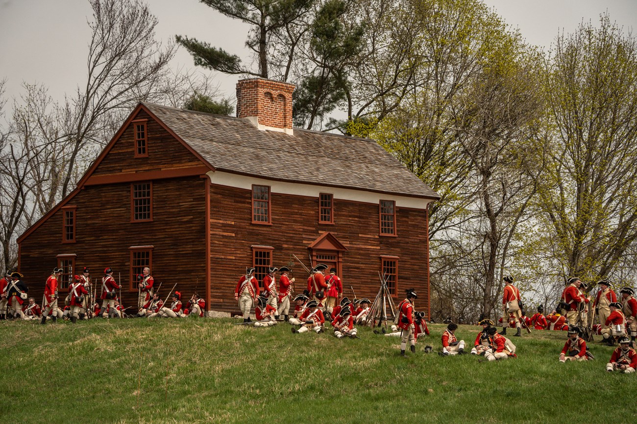 A group of redcoat soldiers sit next to a wooden 18th century house on a hill