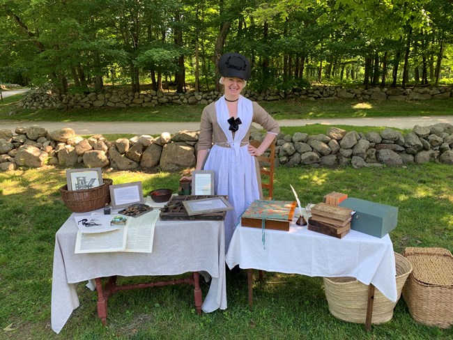 An 18th century woman stands behind a table with materials to print documents