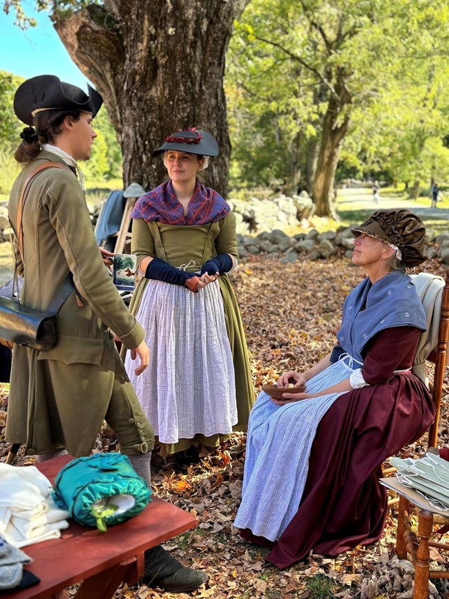 A man in 18th century clothing stands talking to two women in 18th century clothing